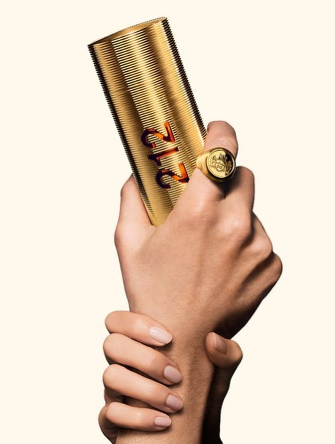 A hand with a gold signet ring holding the gold Carolina Herrera 212 bottle